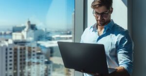 young business professional with laptop standing in front of NYC high rise window