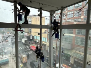 Three rope‑access cleaners hang inside a glass atrium, polishing windows above Manhattan traffic.